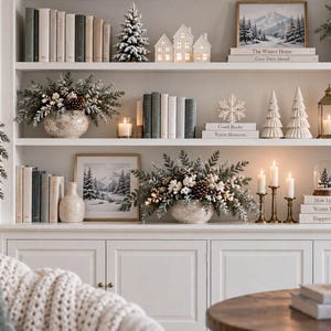 May include: A white built-in shelving unit decorated for winter with books, miniature houses, frosted pine trees, pinecone arrangements, and lit candles. The shelves feature framed winter landscape art and stacked books with titles like The Winter Home.