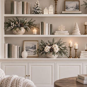 Puede incluir: Una estantería blanca empotrada decorada para el invierno con libros, pequeñas casas de cerámica, un árbol escarchado en miniatura, arreglos de piñas, jarrones blancos, velas en soportes de latón y cuadros de paisajes invernales.