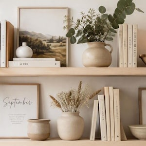 Op de afbeelding: Twee planken van licht hout met decoratieve voorwerpen, waaronder ingelijste landschaps- en botanische kunst, beige keramische vazen, stapels boeken, een houten schaal met een kaars en een hangende groene plant. De tekst September is zichtbaar op een print.