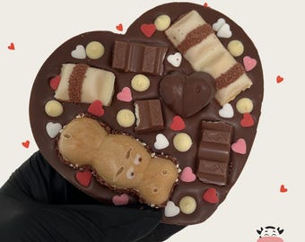 Könnte beinhalten: Ein herzförmiges Schokoladenkonfekt, verziert mit Schokoriegeln, herzförmigen Streuseln und einem Lebkuchenkeks. Die Leckerei wird von einer behandschuhten Hand gehalten, mit einem Logo in der unteren rechten Ecke.