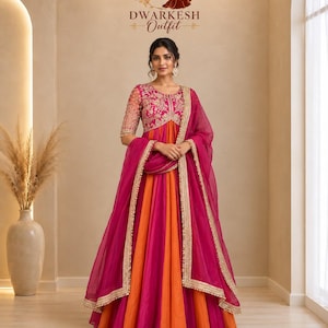 May include: A floor-length anarkali dress featuring alternating panels of orange and magenta fabric. The bodice has three-quarter sleeves with intricate gold embroidery. A matching magenta sheer dupatta with a gold border is draped over the shoulders.