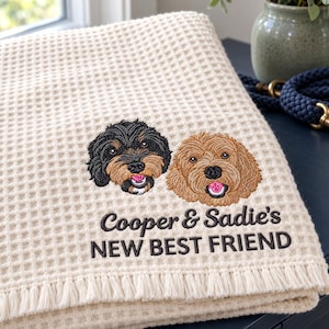 以下が含まれることがあります： フリンジ付きのクリーム色のワッフル織りブランケット。黒とタンの犬とゴールデンブラウンの犬の刺繍ポートレートが施されており、犬の下には黒い糸で「Cooper & Sadie's NEW BEST FRIEND」という文字が刺繍されています。