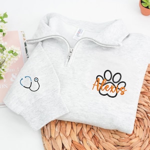 May include: A light gray quarter-zip sweatshirt with a stethoscope embroidered on the left sleeve. The right chest features a paw print design with the name "Alexis" in orange script. The sweatshirt has a zipper closure.