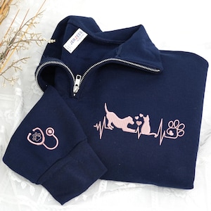 Peut inclure: Sweat-shirt bleu marine à fermeture éclair quart de zip avec un motif brodé rose. Le motif comprend un chien, un chat, une empreinte de patte et un stéthoscope. Le sweat-shirt a une fermeture éclair argentée.