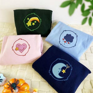 May include: Four folded sweatshirts in dark green, light blue, pink, and navy blue. Each has an embroidered design within a circle. Designs include a shooting star, a cloud with hearts, a heart with a smiling star, and a crescent moon with a star.