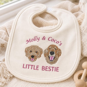 May include: An off-white baby bib featuring embroidered portraits of two dogs. Above the dogs, the text reads "Molly & Coco's" in pink thread. Below the dogs, the text reads "LITTLE BESTIE" in pink thread. The bib has a rounded shape and a finished edge.
