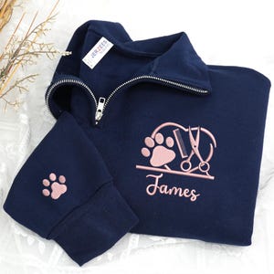 May include: Navy blue quarter-zip sweatshirt with a pink paw print on the sleeve and a larger embroidered design featuring a paw print, scissors, and comb, with the name "James" below.