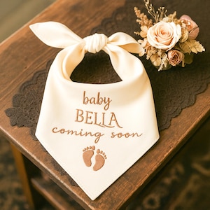 May include: Cream-colored baby bandana with the text "baby BELLA coming soon" embroidered in a brown thread. The bandana features a knotted top and a pair of embroidered baby footprints. A small floral arrangement sits in the background.