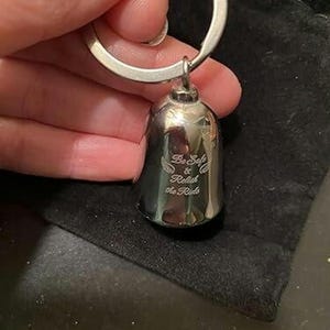 May include: A polished silver-toned metal bell keychain featuring an engraved design with two small wings and the text "Be Safe & Relish the Ride" centered on the front surface. The bell is attached to a circular metal split ring for keys.