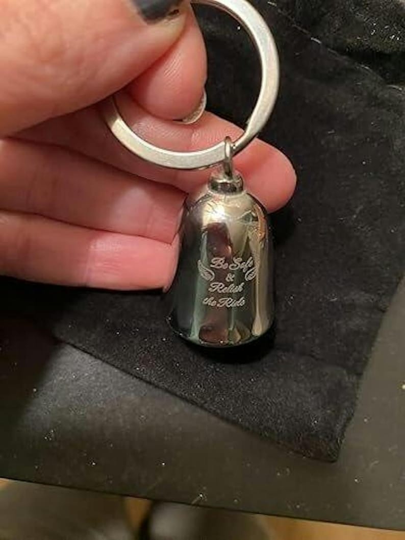 May include: A polished silver-toned metal bell keychain featuring an engraved design with two small wings and the text "Be Safe & Relish the Ride" centered on the front surface. The bell is attached to a circular metal split ring for keys.