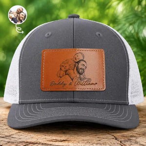 Può includere: Cappellino da camionista grigio con retro in rete bianca e visiera curva. Una toppa in pelle marrone sul davanti presenta un disegno di un uomo e un bambino, con la scritta "Daddy & William". Il cappellino ha una chiusura a scatto.