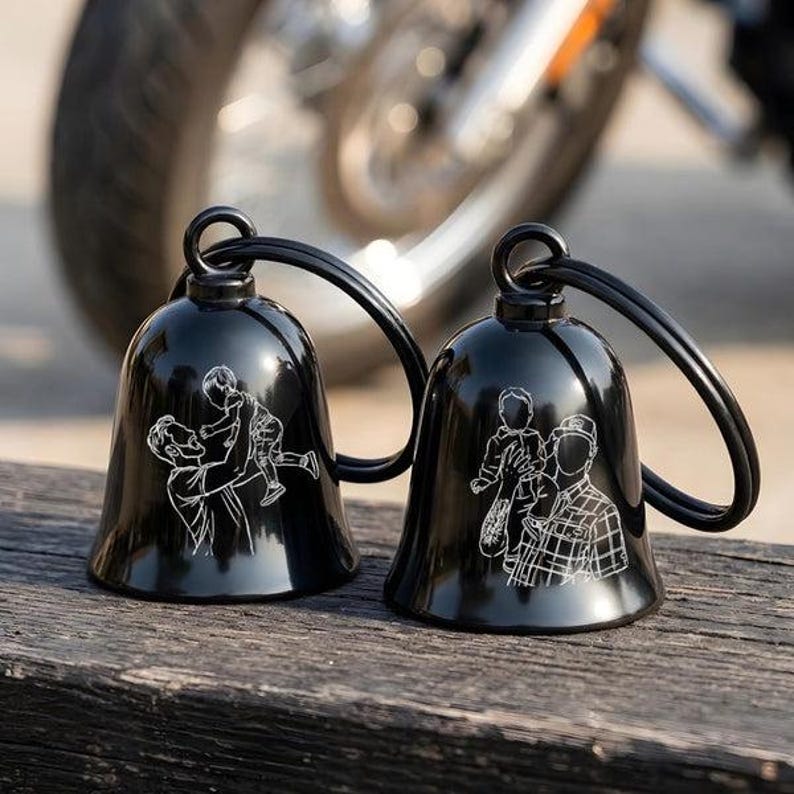 May include: Two glossy black metal bells with attached key rings. Each bell features a white line-art engraving of a person holding a child. The left bell shows a person lifting a child, while the right bell depicts a person wearing a plaid shirt.