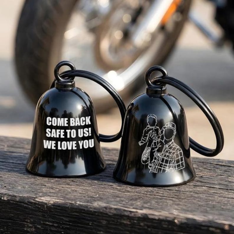 May include: Two glossy black metal bells with attached key rings. One bell features the white text "COME BACK SAFE TO US! WE LOVE YOU". The other bell displays a white line-art illustration of a person holding a small child in their arms.