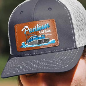 May include: A navy blue trucker hat with a white mesh back. The hat features a brown leather patch with the words "Pontoon Captain" and a graphic of a pontoon boat in blue and gray. The name "William" is also on the patch.