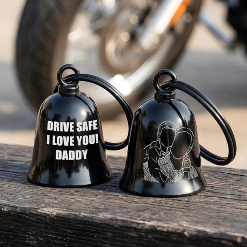 May include: Two glossy black metal bells with attached rings. One bell features the white engraved text DRIVE SAFE I LOVE YOU! DADDY. The other bell displays a white line-art illustration of a person carrying another person on their back.
