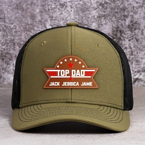 May include: An olive green baseball cap with a black mesh back and a brown leather-like patch on the front. The patch features a red star, white text reading TOP DAD, and the names JACK, JESSICA, and JAME printed in white on a brown background.
