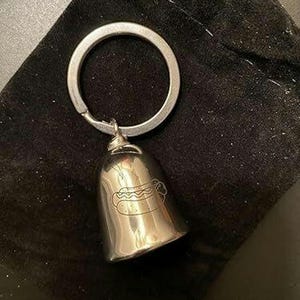 May include: A silver-toned metal bell keychain featuring a simple line drawing of a hot dog engraved on the front surface. The bell is attached to a circular split ring, providing a functional accessory for keys or bags with a polished finish.