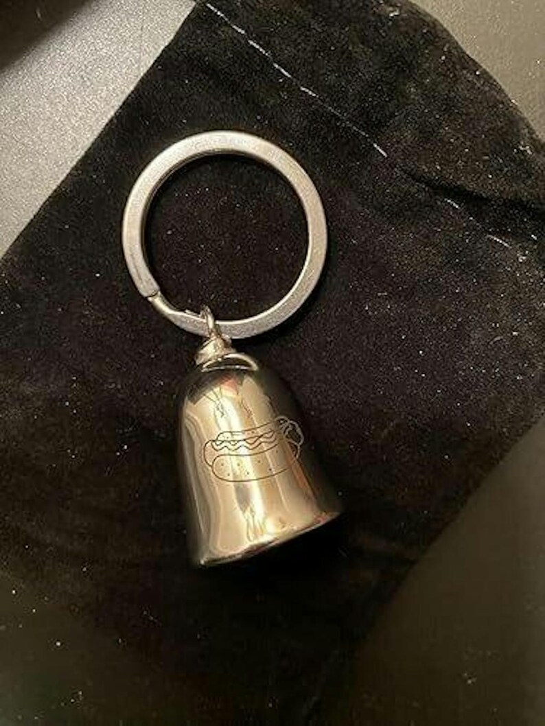 May include: A silver-toned metal bell keychain featuring a simple line drawing of a hot dog engraved on the front surface. The bell is attached to a circular split ring, providing a functional accessory for keys or bags with a polished finish.