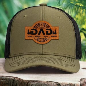 Peut inclure: Casquette de camionneur vert olive et noire avec un écusson en cuir personnalisé. L'écusson porte l'inscription "DAD" avec le texte "THE MAN + THE MYTH + THE LEGEND" et les noms Sophia, Marcus, Amelia et Jackson.