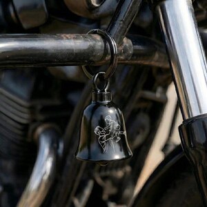 May include: A glossy black metal bell attached to a circular ring. The bell features a white line-art engraving of an adult lifting a small child into the air. The object is suspended from a dark metal frame with cylindrical components.