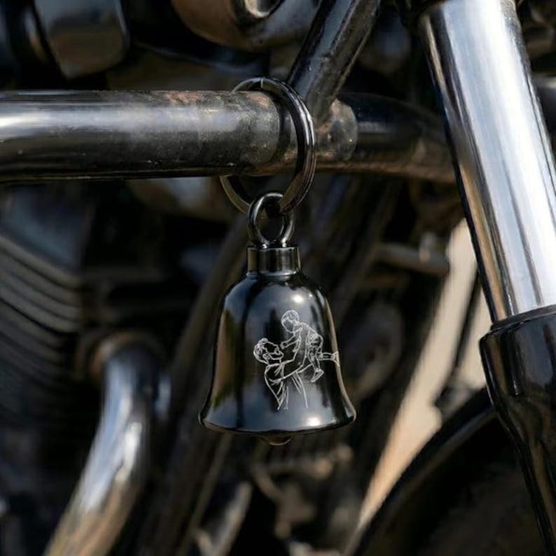 May include: A glossy black metal bell attached to a circular ring. The bell features a white line-art engraving of an adult lifting a small child into the air. The object is suspended from a dark metal frame with cylindrical components.