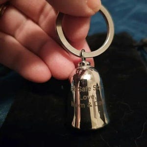 May include: A silver-toned metal bell keychain featuring a circular split ring. The bell has an engraved inscription that reads: Be Safe Son, Love, Mom & Dad. The surface of the bell is polished and reflective, showing a smooth, metallic finish.