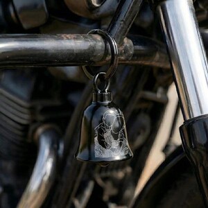 May include: A black metal bell keychain featuring a white laser-engraved line art illustration of a person embracing another person. The bell is attached to a circular metal ring and hangs from a black motorcycle frame component.