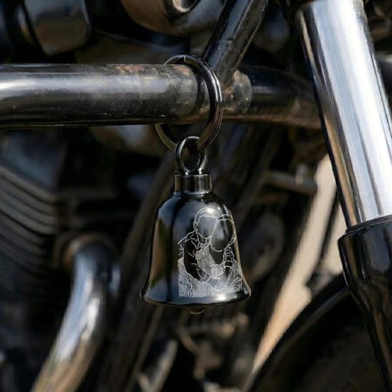May include: A black metal bell keychain featuring a white laser-engraved line art illustration of a person embracing another person. The bell is attached to a circular metal ring and hangs from a black motorcycle frame component.