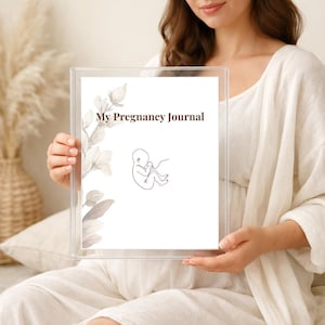 Könnte beinhalten: Ein klarer Acrylrahmen hält eine weiße Seite mit dem Text "My Pregnancy Journal". Die Seite zeigt eine Strichzeichnung eines Fötus und florale Akzente. Das Journal wird von einer Person in einem weißen Outfit gehalten.