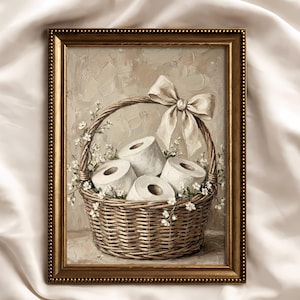 Puede incluir: Pintura enmarcada que representa una cesta de mimbre llena de rollos de papel higiénico blancos. La cesta está adornada con pequeñas flores blancas y un gran lazo color crema. El marco es dorado.