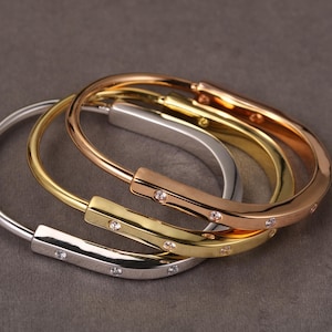 May include: Three stylish bangles in silver, gold, and rose gold. Each bracelet has a line of small, sparkling gemstones. The bangles are stacked, highlighting their different metallic finishes and design variations.