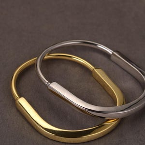 May include: Two stylish bangles, one gold and one silver. Each has a smooth, curved shape. The bracelets are set against a dark grey backdrop, which accentuates their metallic finish. These accessories are ideal for adding a touch of elegance to any outfit.