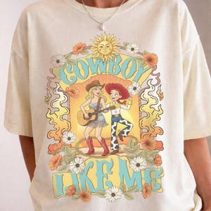 May include: A cream-colored short-sleeve t-shirt featuring a graphic print of two characters, one playing a guitar, surrounded by flowers, a sun, and flames. The text "COWBOY LIKE ME" is printed in large, light blue letters above and below the illustration.