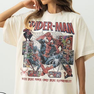 May include: Ivory-colored t-shirt featuring a Spider-Man graphic. The design includes the words "Spider-Man" in red, along with comic book illustrations of the superhero. The text "WITH GREAT POWER COMES GREAT RESPONSIBILITY" is also present.