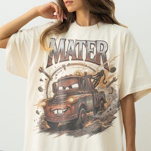 May include: Ivory-colored t-shirt featuring a graphic of Mater from the movie Cars. The design includes the word "MATER" above the truck, with a background of tools and a speedometer. The shirt is oversized.
