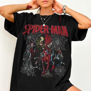 Pu&ograve; includere: T-shirt nero con grafica di Spider-Man e la scritta rossa "SPIDER-MAN". La grafica include vari personaggi di Spider-Man e un disegno a ragnatela. La maglietta &egrave; in stile casual e oversize.