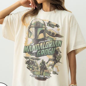 May include: Ivory-colored oversized t-shirt featuring a graphic from the Star Wars series, "The Mandalorian and Grogu." The design includes images of the Mandalorian, Grogu, and spacecraft, with the phrase "This is the Way."