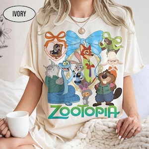 Puede incluir: Camiseta de color marfil con un gr&aacute;fico colorido de personajes de la pel&iacute;cula animada "Zootopia". El dise&ntilde;o incluye un lazo azul, un marco en forma de coraz&oacute;n y la palabra "Zootopia" en verde. La camiseta tiene mangas cortas.