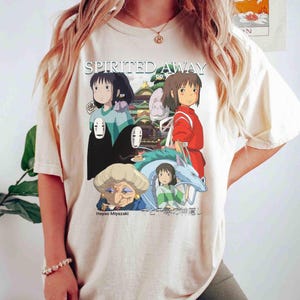 Pode incluir: Camiseta creme com um gr&aacute;fico do filme de anima&ccedil;&atilde;o "A Viagem de Chihiro". O design inclui personagens e o t&iacute;tulo do filme em branco. A parte inferior do gr&aacute;fico inclui o texto "Hayao Miyazaki" e texto japon&ecirc;s.