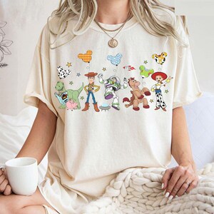 Puede incluir: Camiseta color crema con un gr&aacute;fico colorido de personajes animados y globos. El dise&ntilde;o incluye un dinosaurio, un vaquero, un astronauta, un caballo y una vaquera. La camiseta es de estilo casual y oversize.