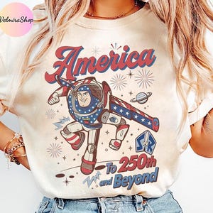May include: Cream-colored t-shirt featuring a cartoon illustration of a space ranger in patriotic colors. The design includes the word "America" in red and blue script, with the text "To 250th and Beyond" below.