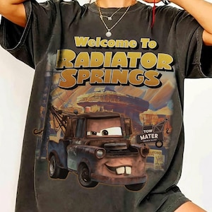 Puede incluir: Camiseta gris oscuro con el texto "Welcome To RADIATOR SPRINGS" en dorado y blanco. El dise&ntilde;o incluye a Mate de Cars. La camiseta tiene un aspecto vintage y desgastado.