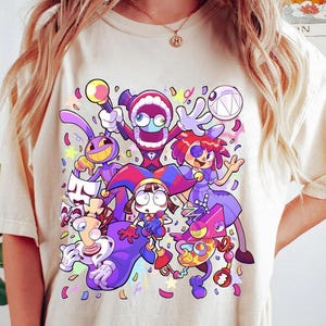 May include: A cream-colored t-shirt featuring a vibrant, multi-colored graphic print of various cartoon characters. The design includes figures with exaggerated features, bright purple, red, and yellow accents, and scattered confetti shapes.