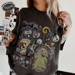 Puede incluir: Una camiseta de manga corta gris carb&oacute;n con un estampado gr&aacute;fico de personajes de Pesadilla antes de Navidad, incluidos Jack Skellington, Sally y Oogie Boogie, junto con iconos como un lazo, telara&ntilde;as y dulces de ma&iacute;z.