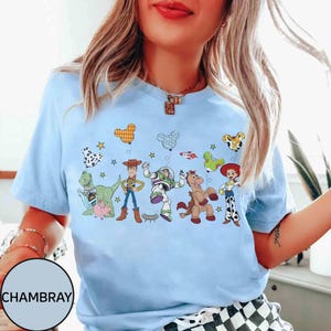 Puede incluir: Camiseta azul claro con un gr&aacute;fico colorido de personajes de Toy Story y globos. El dise&ntilde;o incluye a Woody, Buzz Lightyear, Jessie y otros personajes. La palabra "CHAMBRAY" est&aacute; impresa en la parte inferior izquierda.