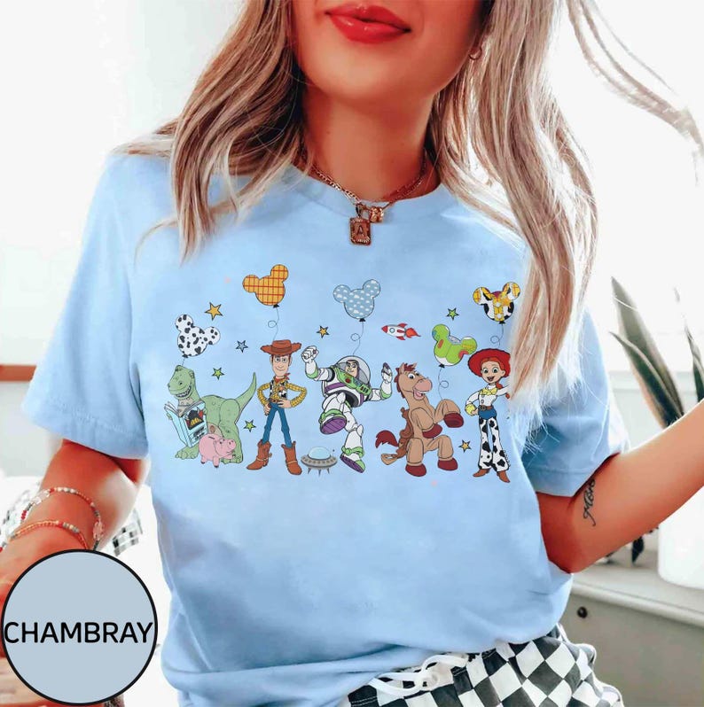 Puede incluir: Camiseta azul claro con un gr&aacute;fico colorido de personajes de Toy Story y globos. El dise&ntilde;o incluye a Woody, Buzz Lightyear, Jessie y otros personajes. La palabra "CHAMBRAY" est&aacute; impresa en la parte inferior izquierda.