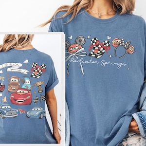 May include: A light blue t-shirt featuring a graphic of the Cars movie characters and the text "Radiator Springs". The design includes images of race cars, checkered flags, and Mickey Mouse ears.
