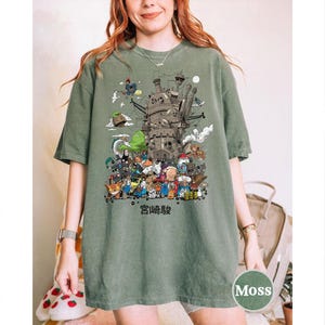 以下が含まれることがあります： モスグリーンのTシャツ。城と様々なキャラクターの詳細なイラストが描かれています。下部には日本語のテキストがあります。ゆったりとしたフィット感のシャツです。