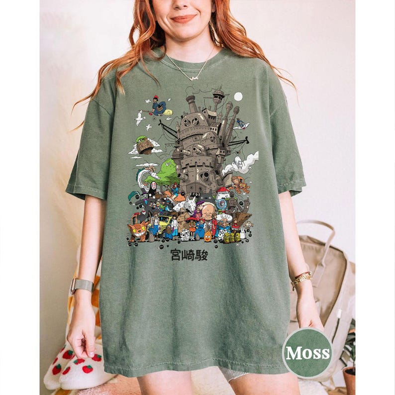 以下が含まれることがあります： モスグリーンのTシャツ。城と様々なキャラクターの詳細なイラストが描かれています。下部には日本語のテキストがあります。ゆったりとしたフィット感のシャツです。
