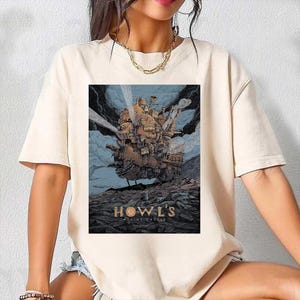 Puede incluir: Camiseta color crema con una ilustraci&oacute;n detallada del Castillo Ambulante de la pel&iacute;cula animada. La obra de arte representa el castillo sobre un fondo de cielos azules y nubes oscuras, con el texto "HOWL'S MOVING CASTLE" en la parte inferior.